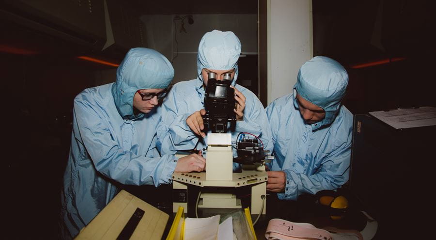 Three electronics students with lab coats using a microscope