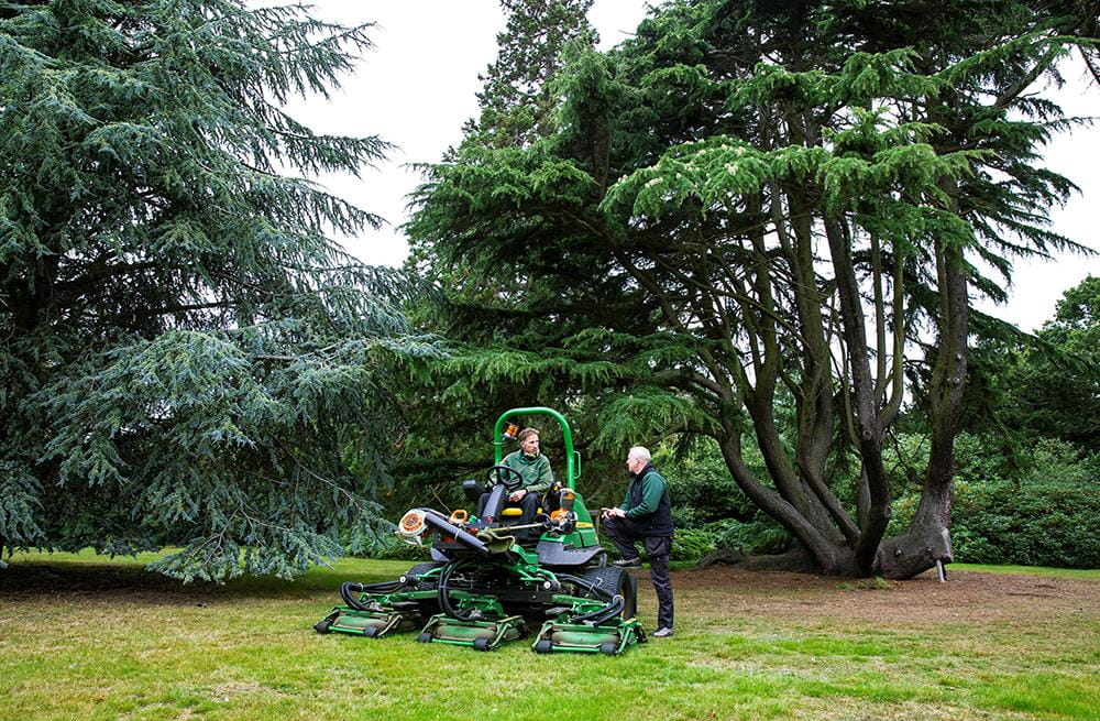 Two members of the Grounds team discuss as one sits on a lawnmower in front of a mixture of old trees.