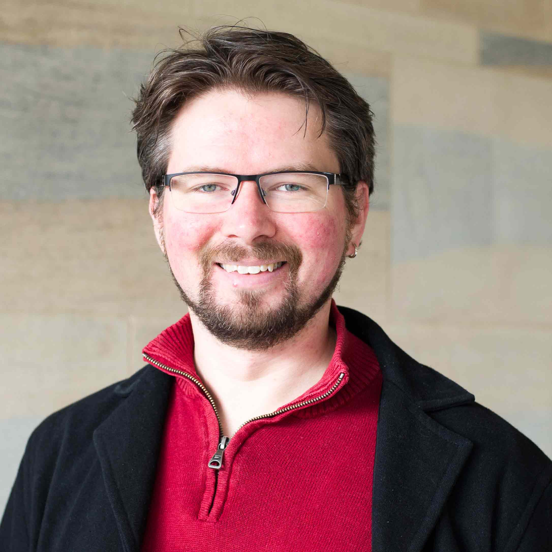 Dr Liam Jarvis, smiling, wearing glasses, and wearing a red top under a black jacket