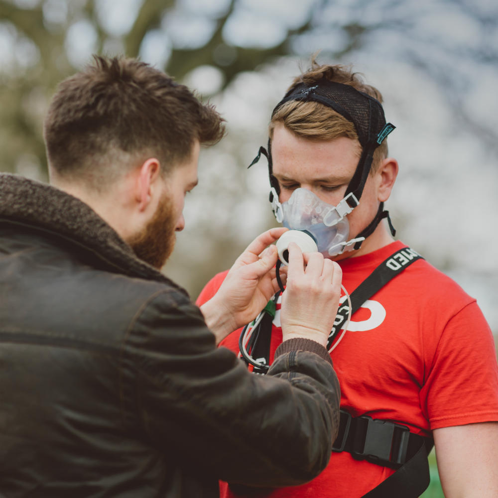 A researcher adjusts an oxygen mask worn by a person in a red t-shirt, who has some other equipment strapped around his chest.